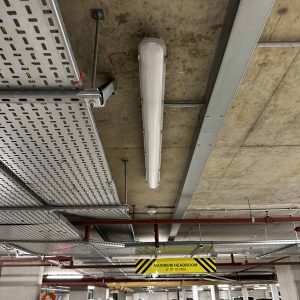 A ceiling with industrial lighting, electrical conduit, and safety signage in a modern parking garage.